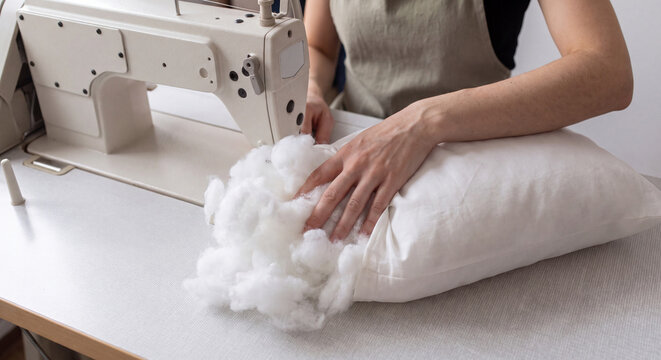 A female artisan filling a white fabric pillowcase with soft polyester fiber stuffing while sitting at a sewing machine workstation