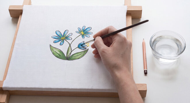 Overhead view of an illustrator holding a fine brush and applying colorful layers to a botanical flower painting on an angled wooden desk board