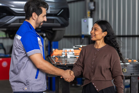 Mechanic shakes hands with customer in EV repair workshop beside electric car and battery pack, showing service agreement, maintenance trust, customer support, clean energy and automotive aftercare.