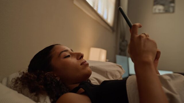Multiethnic young woman lying in bed, browsing smartphone before setting device down thoughtfully. Footage fits mental wellness campaigns, insomnia awareness projects, or digital detox ads