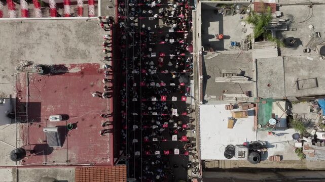 Straight down view of concrete rooftops and a street lined with red and white paper flags in San Juan de Ocotan, Jalisco, as horses and costumed riders move through the waiting crowd. Aerial top down