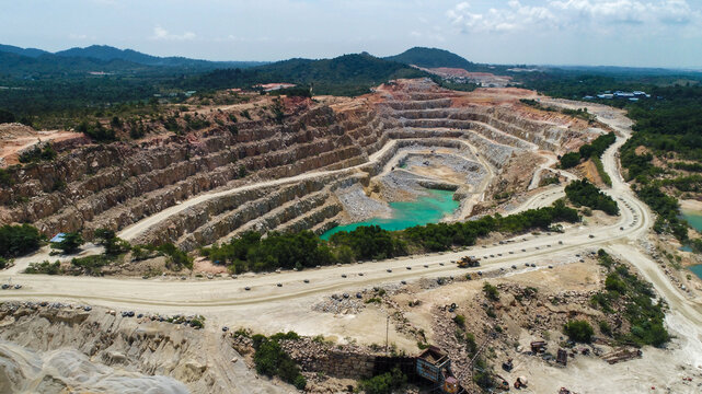 Aerial view of a large open-pit gravel mine with terraced walls, turquoise water at the bottom, and heavy machinery on the surrounding dirt roads in Karimun, Kepulauan Riau, Indonesia.