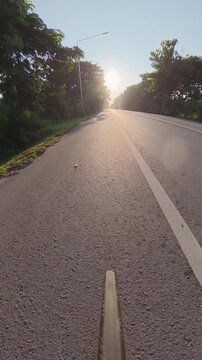 Cyclist POV riding on a quiet rural road toward low golden sunlight, with tree-lined surroundings, long shadows and an empty asphalt roadway.
