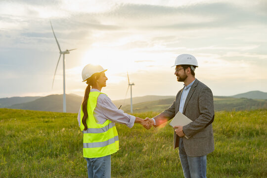 Two engineers in hard hats and safety vests shake hands in a field with wind turbines in the background during sunset