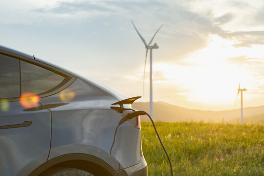 An electric vehicle is plugged into a charging station amidst a field with wind turbines in the background during sunset