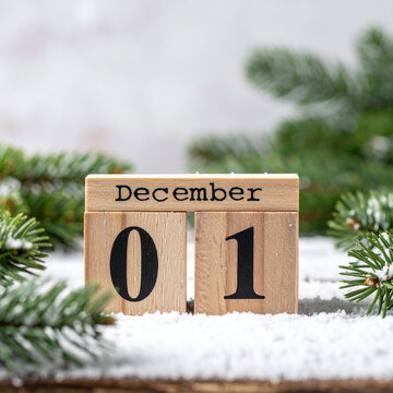 Wooden calendar cubes with text DECEMBER 1 on a snow-covered rustic surface, fresh fir tree branches, cozy festive atmosphere. Close-up.