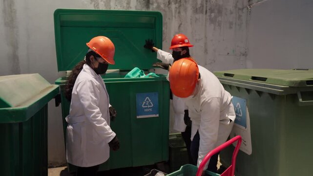 Three workers at a recycling plant deposit empty High-Density Polyethylene (HDPE) containers into a collection bin, contributing to a sustainable construction project by upcycling plastic waste into e
