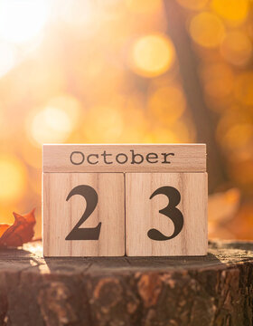 Wooden calendar cubes with text OCTOBER 23 on a rustic wooden pedestal, warm orange autumn leaves, soft bokeh on background, golden seasonal sunlight.