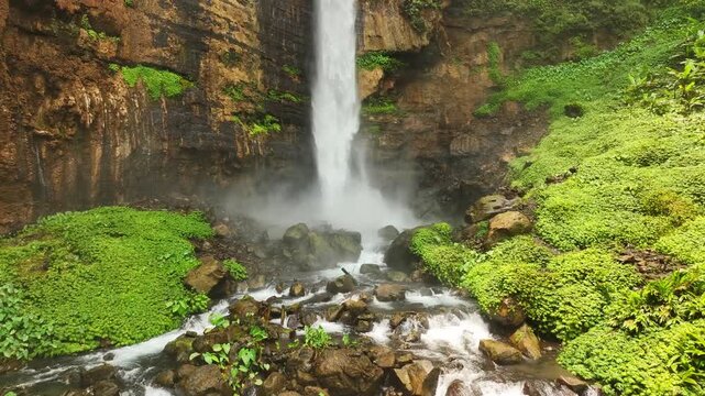 Aerial view of the cascading Kapas Biru Waterfall surrounded by lush green vegetation and rocky cliffs in Kecamatan Pronojiwo, Jawa Timur, Indonesia.