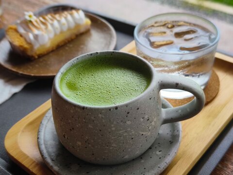 Hot matcha green tea served in a speckled ceramic cup on a wooden tray alongside a glass of iced water and a slice of lemon meringue pie