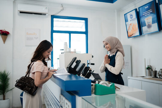 Young customer and shop attendant share friendly moment at bright dessert counter in modern cafe, showing payment area, menu boards, product displays, and blue accents in welcoming retail interior.