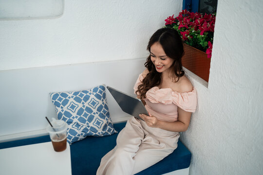Young woman in peach off shoulder outfit sits on blue bench, smiling while reading tablet beside iced drink, patterned cushion, and vivid red flowers in cozy white cafe nook with soft natural light