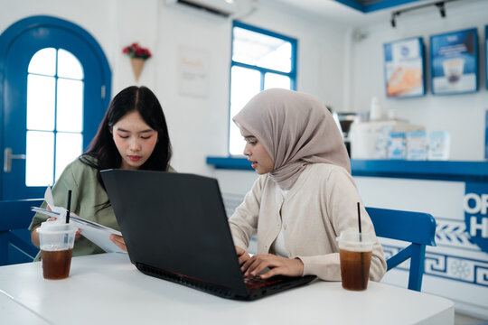 Two young women sit at white table, reviewing papers and typing on laptop inside bright blue cafe interior, with iced drinks nearby, showing focused study, planning, and informal teamwork.