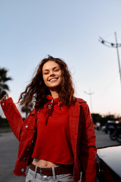 Young woman in red jacket smiling outdoors. Beautiful girl with curly hair enjoying warm sunlight. Urban street lifestyle portrait of happy female.