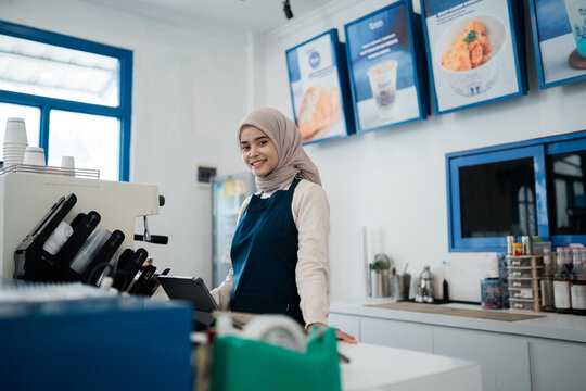 Young woman in hijab smiles behind cafe counter, standing beside espresso machine, takeaway cups, payment device, and neatly arranged drink station inside bright modern coffee shop interior.