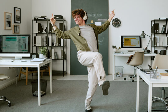 Young adult Caucasian man wearing headphones dancing in modern office celebrating career success. Image suitable for corporate wellness campaigns, workplace happiness, mental wellbeing ads