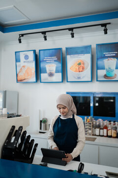Young woman in hijab at cafe counter typing on tablet while blue menu boards, drink bottles, and service tools frame clean bright workspace in modern coffee shop interior.