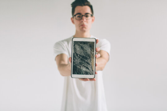 Nerd is wearing glasses. Student presenting a broken black tablet behind glass. upset man holds a out-of-use tablet or smartphone. Isolated on a light background. Broken screen