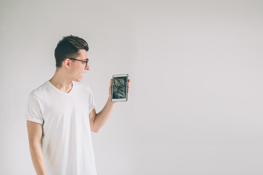 Nerd is wearing glasses. Student presenting a broken black tablet behind glass. upset man holds a out-of-use tablet or smartphone. Isolated on a light background. Broken screen
