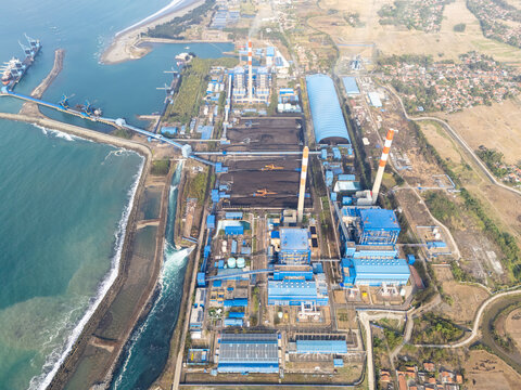Aerial view of the coal-fired power plant with large smokestacks, coal storage piles, and conveyor belts situated along the coastline in Cilacap Selatan, Central Java, Indonesia.