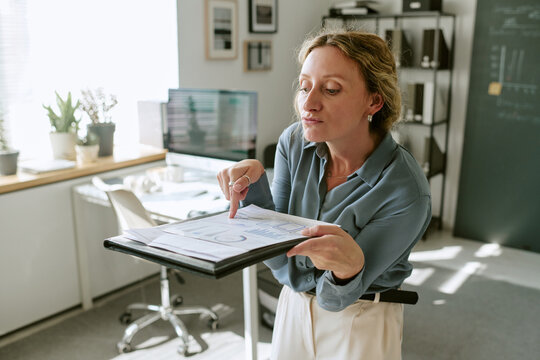 Middle aged Caucasian woman pointing at financial chart on folder in modern office. Concept of data analytics and business strategy, suitable for corporate marketing