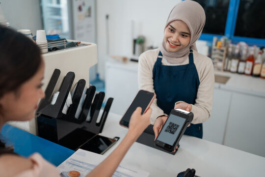 Friendly store employee in hijab accepts mobile payment from customer at bright retail counter, showing secure checkout with smartphone scan, card terminal, and casual service interaction.