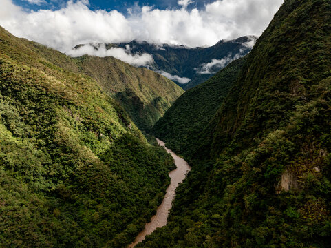 Aerial view of the Urubamba River winding through a deep, lush green valley surrounded by steep Andean mountains under a cloudy sky in Aguas Calientes, Cusco, Peru.