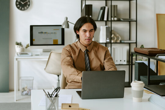 Young adult man working on laptop in modern office, typing reports and analyzing data. Image suitable for business communication, corporate workflow, technology marketing