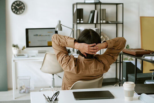 Rear view of young adult businessman relaxing with hands behind head at desk. Ideal for corporate mental wellbeing, workplace stress relief and HR campaigns