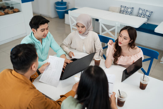 Small group of young adults reviews documents and laptop together in bright casual cafe setting, exchanging ideas over drinks, with collaborative mood, modern seating, and productive teamwork.