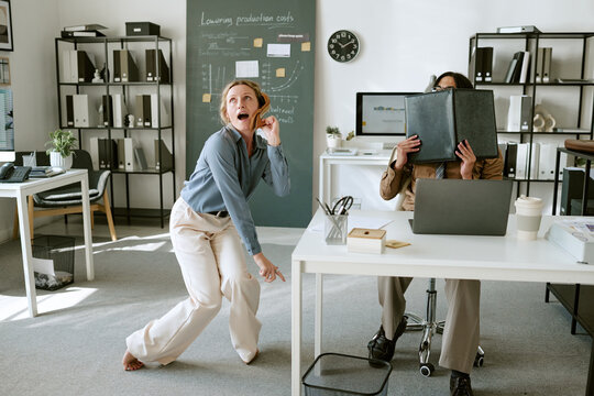 Playful Caucasian young adult woman dancing in office while colleague hides behind folder. Useful for workplace wellness, stress relief and corporate culture marketing campaigns