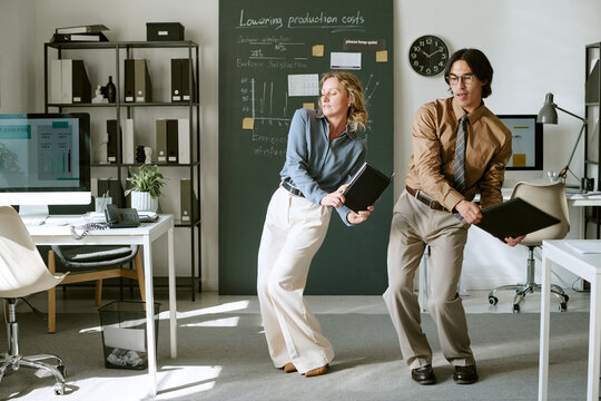 Caucasian middle aged businesswoman and young adult businessman dancing playfully with folders in office, celebrating success, perfect for corporate culture campaigns
