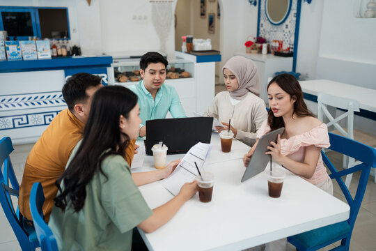 Five young adults discuss work together at white cafe table, using laptop, tablet, notes, and iced drinks in bright modern interior, showing teamwork, planning, and friendly business collaboration.