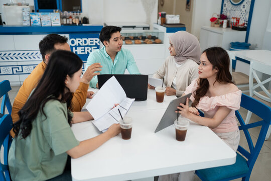 Group of young professionals talking at cafe table with laptop, papers, tablet, and iced drinks, showing casual teamwork, brainstorming, planning, and modern startup collaboration in bright interior