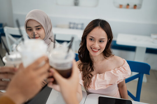 Two young women sit together at desk, smiling while friends in foreground raise iced drinks toward them. Bright modern room, soft daylight, casual conversation, warm social atmosphere.
