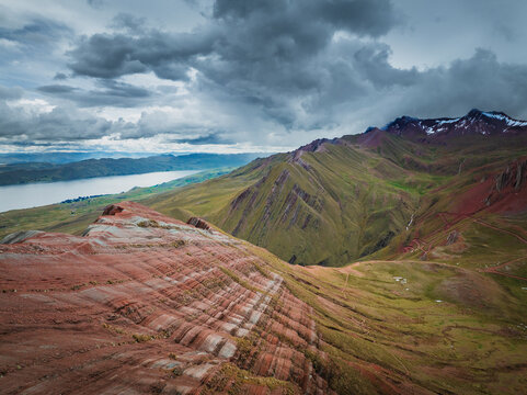 Aerial view of the striated rock formations of Pallay Punchu Rainbow Mountain with green slopes and Langui Layo lake under a dramatic cloudy sky in Layo, Cuzco, Peru.
