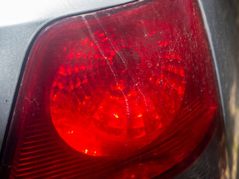 A detailed close-up shot of a dusty and scratched red rear taillight assembly on a grey passenger vehicle. The image highlights the texture of dry mud streaks, fine cracks, and dust settled on the glo