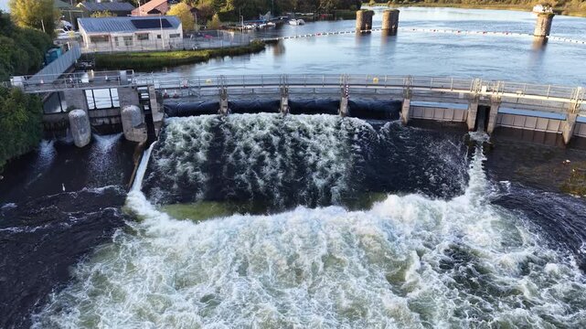Aerial view of Salmon Weir gates releasing turbulent water into the River Corrib in Galway City, Ireland, with stone structures and the upper river behind.
