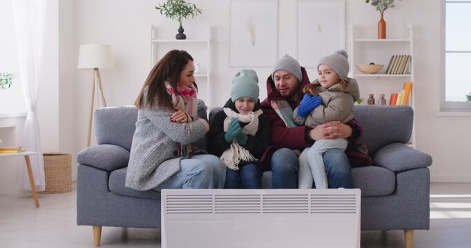 People freezing at home during winter. Family trying to warm up in very cold apartment. Young mom, dad and children in warm coats, jackets, hats and gloves together sitting on sofa by electric heater