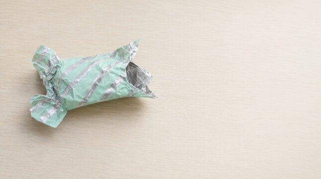 Empty crumpled chewing gum wrapper made of shiny silver and green foil on light wooden table, discarded trash and recycling concept.