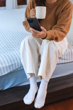 Vertical closeup image of a woman holding and using smart phone while drinking coffee on the bed at home