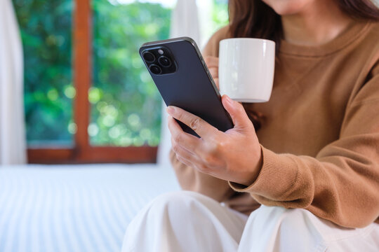 Closeup image of a woman holding and using smart phone while drinking coffee on the bed at home