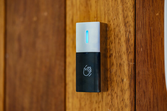 Close up view of a sleek touchless door release sensor with blue light indicator and wave hand icon installed on a natural vertical wood surface