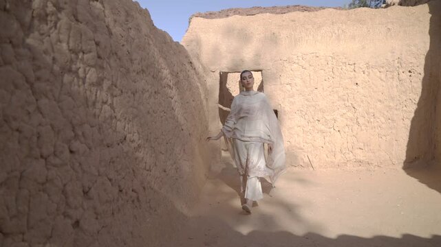 Tracking shot of an elegant woman in a white embroidered dress walking through a rustic mud brick alley in AlUla