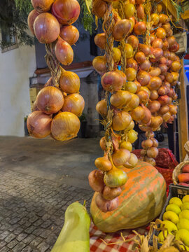 Fresh braided strings of yellow and red onions hanging outdoors at a vibrant local farmers market alongside giant pumpkins and fresh vegetables