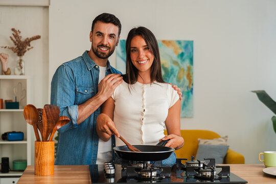 A smiling couple shares a lively kitchen moment as one stirs a pan while the other seasons. The close up composition highlights hands and utensils, creating a warm, inviting focus on home cooking now