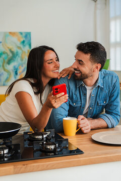 A smiling couple leans on a wooden kitchen island, sharing a moment over a bright red phone. They look closely, exchanging warm expressions as a mug and plate sit nearby. Their calm vibe feels warm