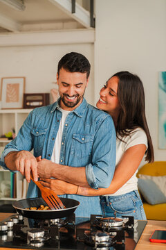 Two people laugh while cooking together at a compact stove, one stirs vegetables with a wooden spatula, camera captures cheerful hands and a warm home mood. Close crop highlights motion and smiles.