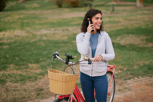 Young woman enjoying an active healthy lifestyle, smiling while talking on a mobile phone, standing with her bicycle in a green park, staying connected outdoors