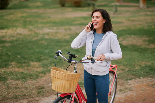 Woman smiling and laughing while talking on a mobile phone, enjoying a leisure time outdoor activity with her bicycle in a park, communicating joyfully